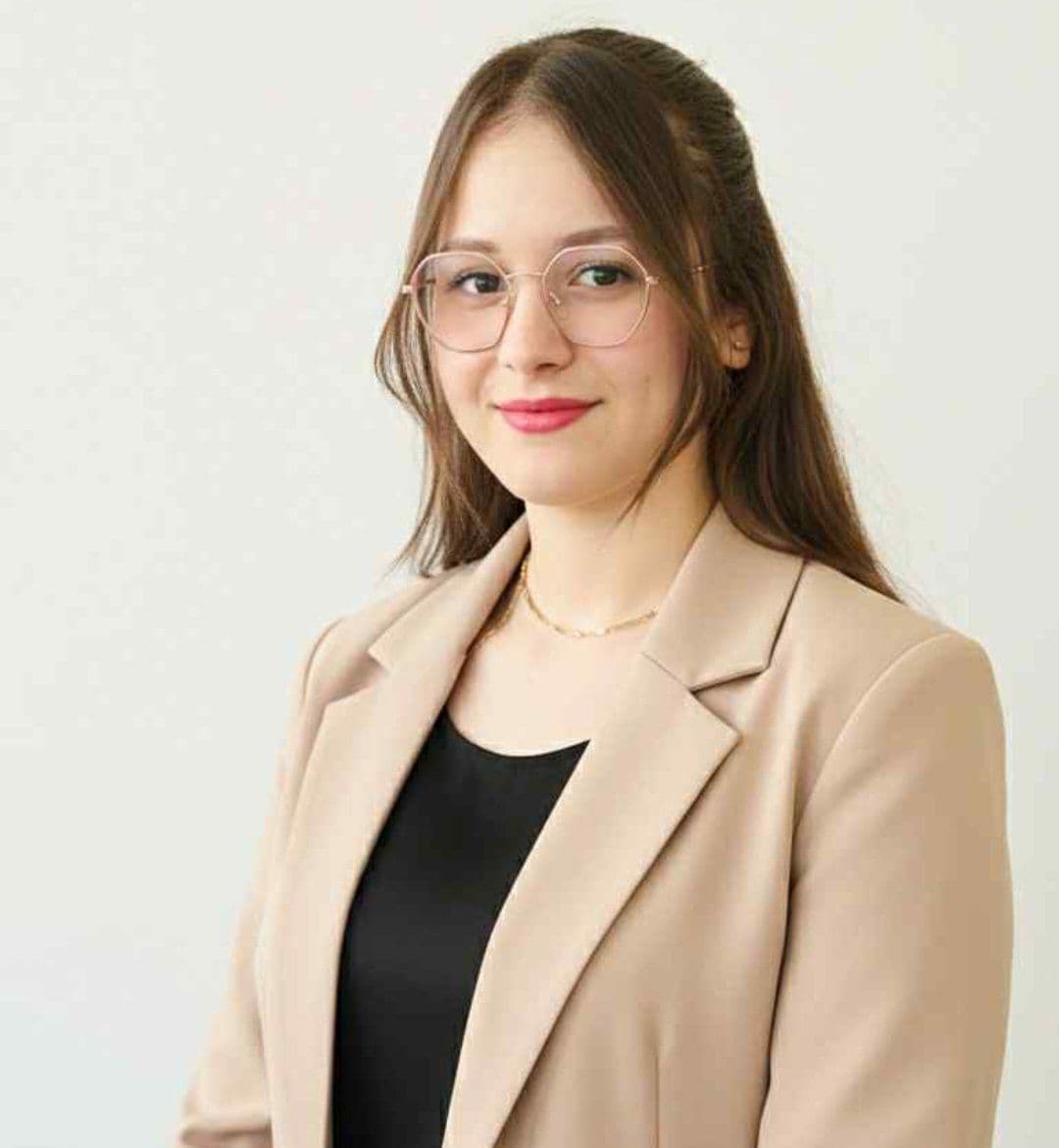 Junge Frau mit Brille, braunen Haaren, beigem Blazer und schwarzem Oberteil vor weißem Hintergrund.