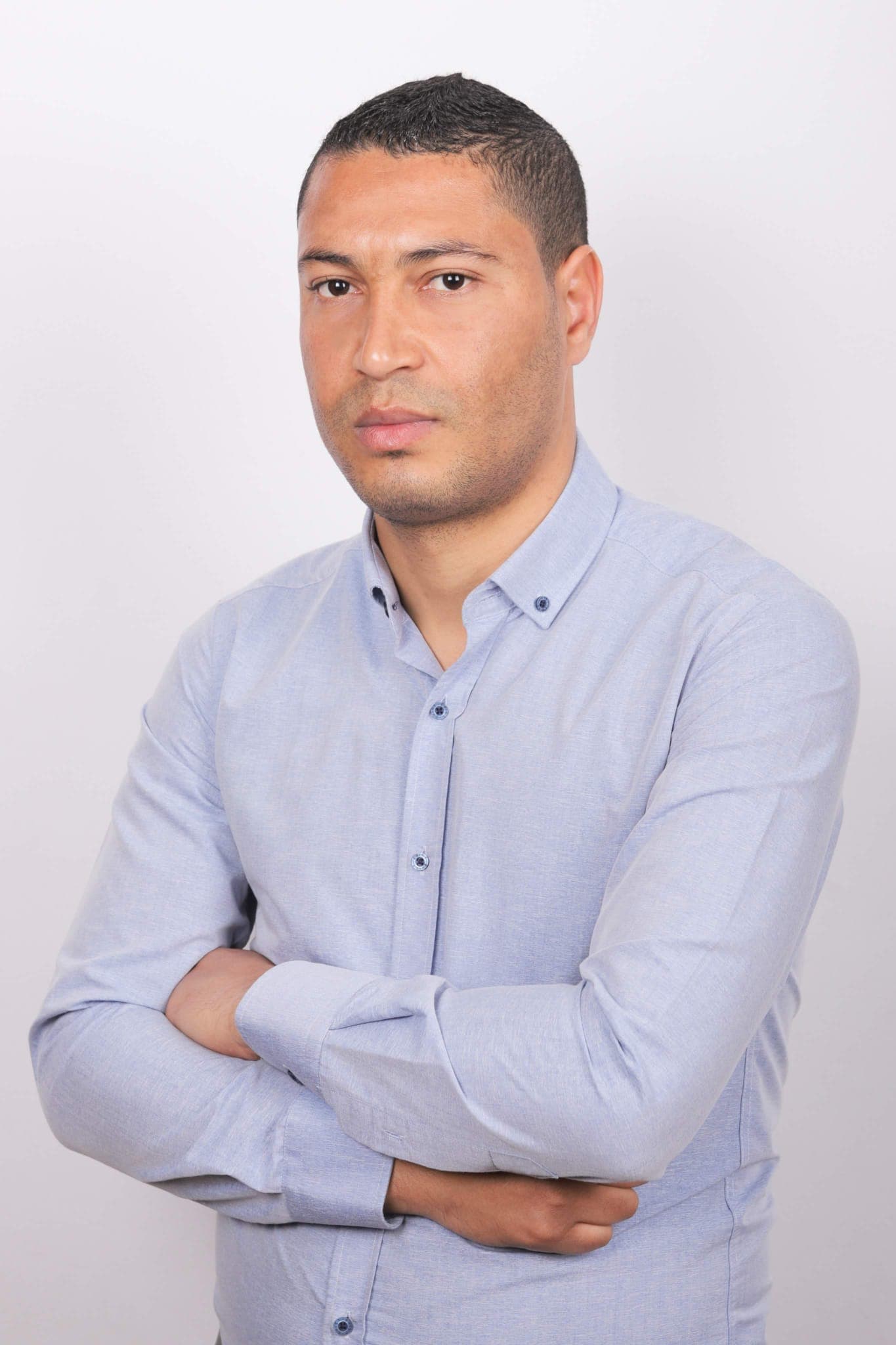 Ein Mann mit verschränkten Armen trägt ein hellblaues Hemd vor einem schlichten weißen Hintergrund.