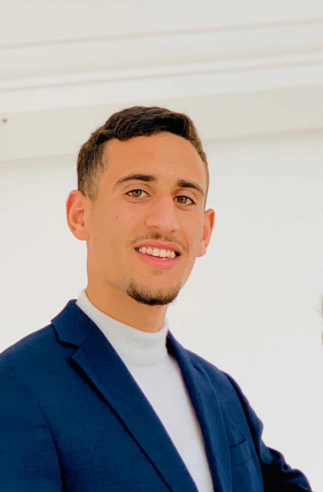 Lächelnder junger Mann mit kurzem Bart, blauem Sakko und weißem Rollkragenpullover vor hellem Hintergrund.