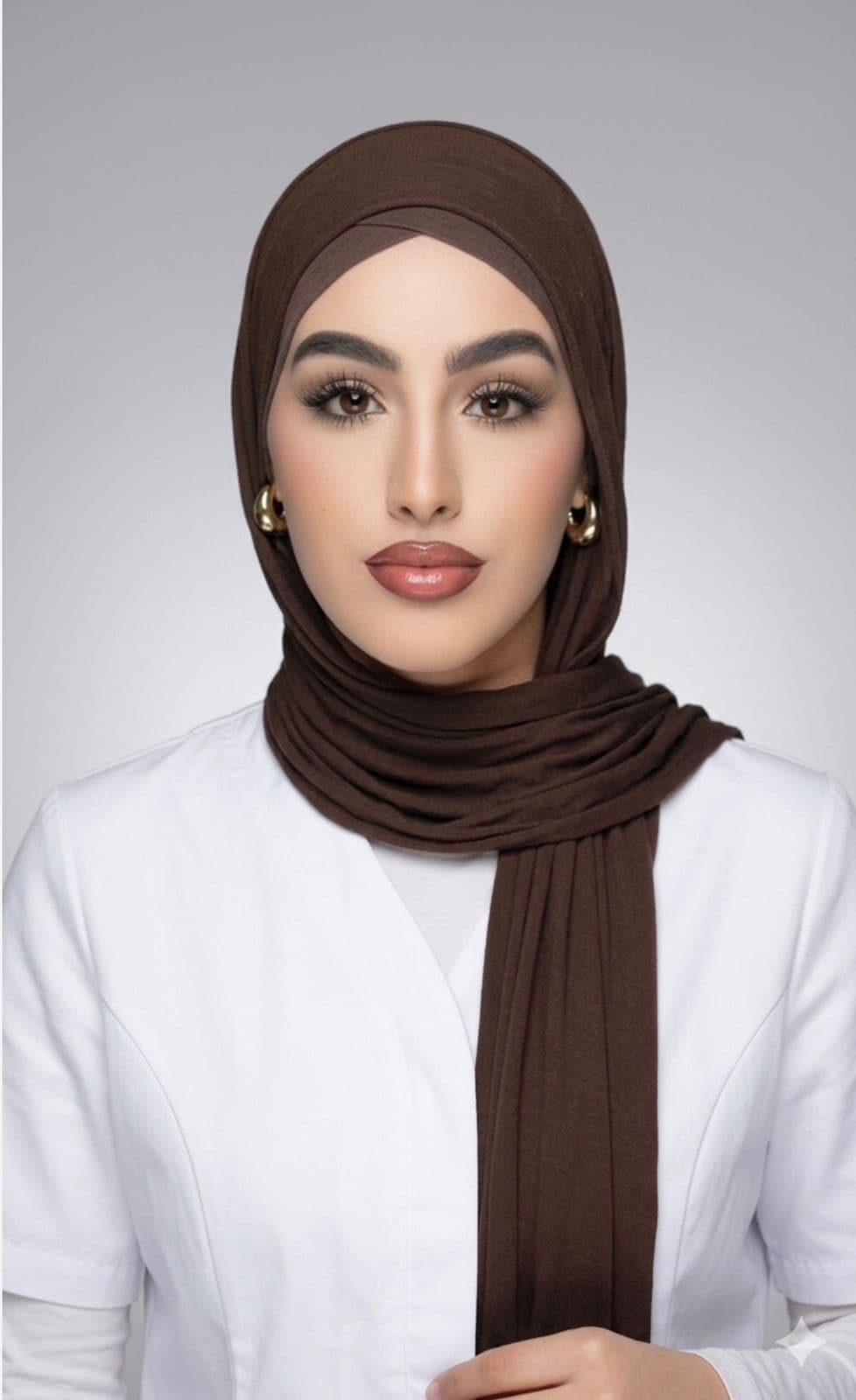 Frau mit braunem Hidschab, weißem Oberteil, elegantem Make-up und goldenen Ohrringen vor grauem Hintergrund.